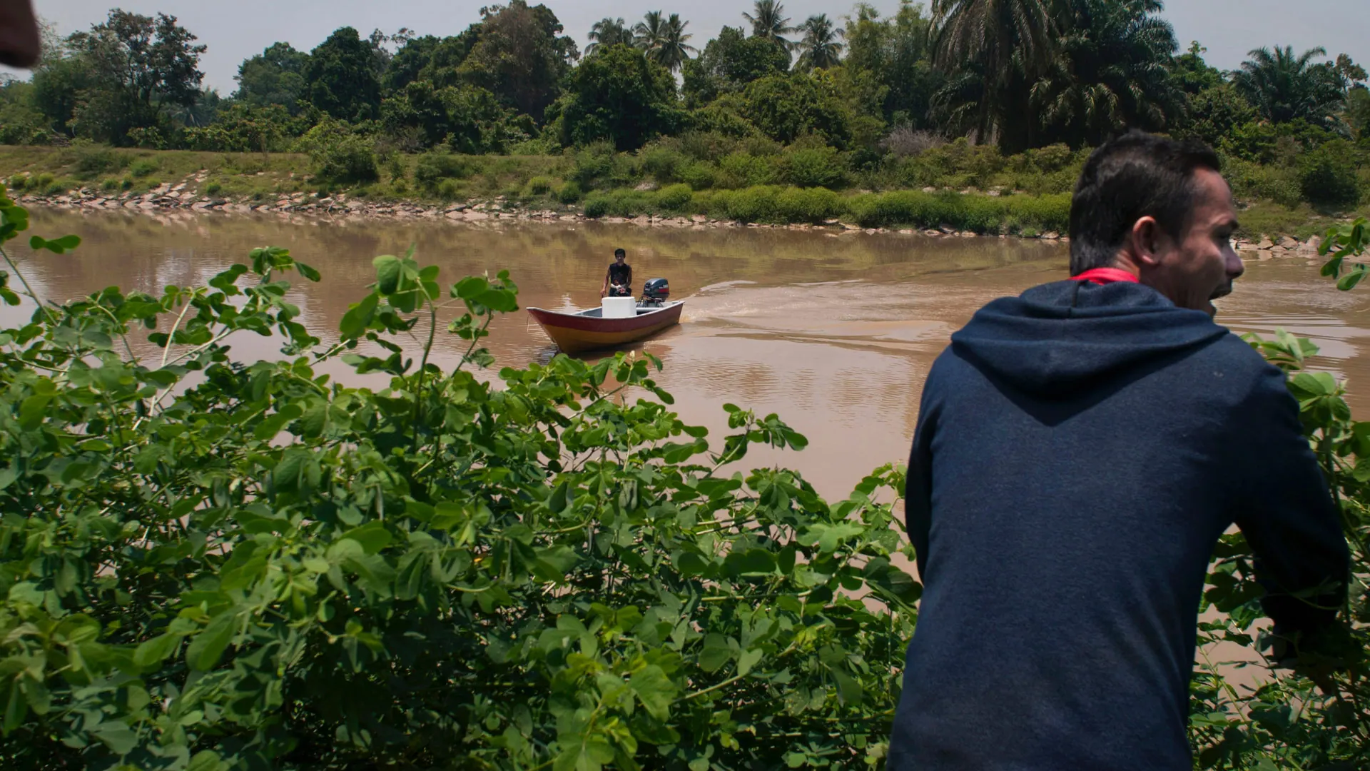 A man in a hooded top watches a small wooden boat cross a wide brown river at the Thailand to Malaysia border, 2012.