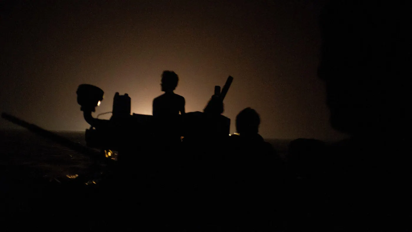 Silhouettes of people boarding a small boat at night, lit only by a distant orange glow, 2012.