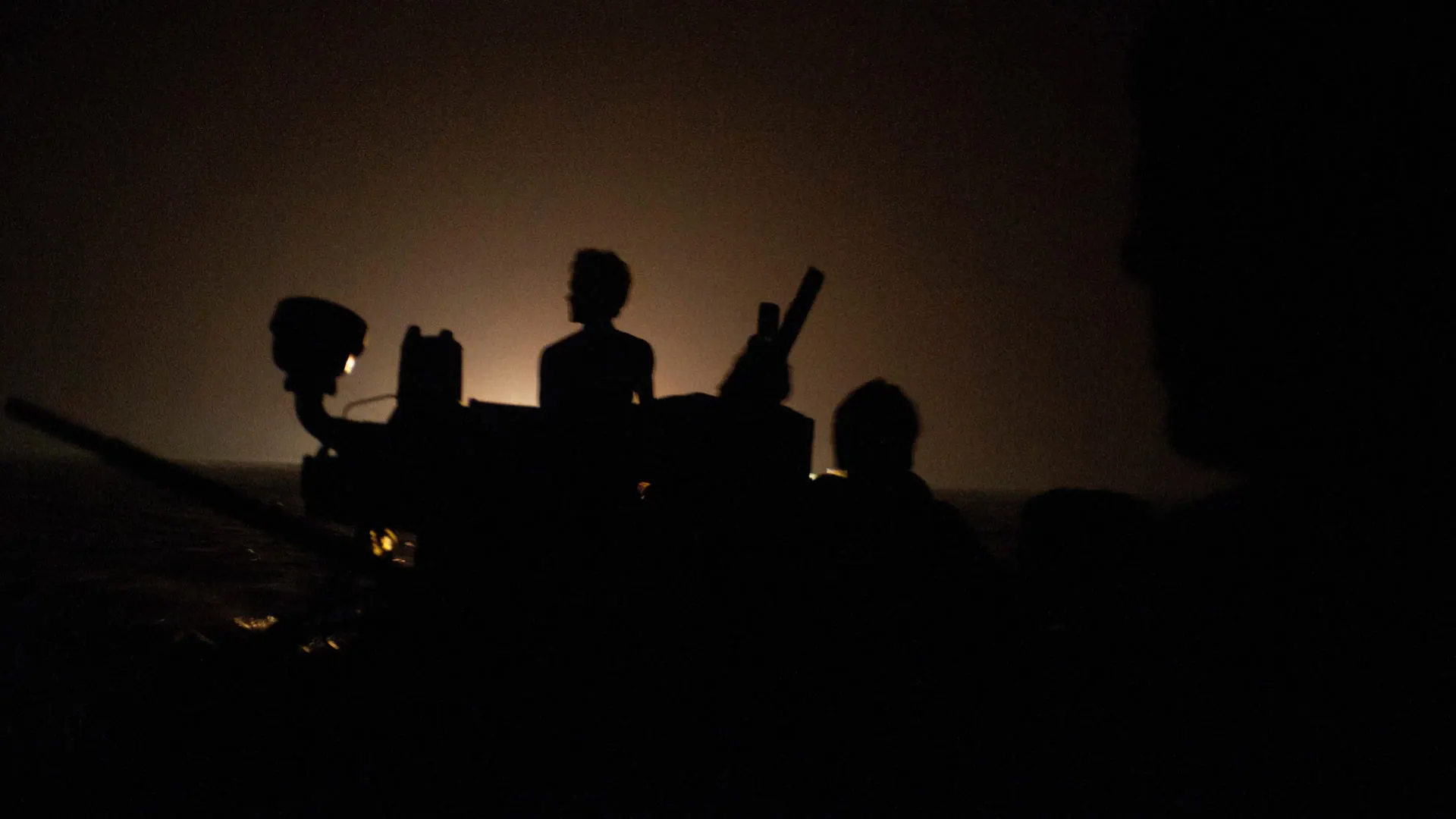 Silhouettes of people boarding a small boat at night, lit only by a distant orange glow, 2012.