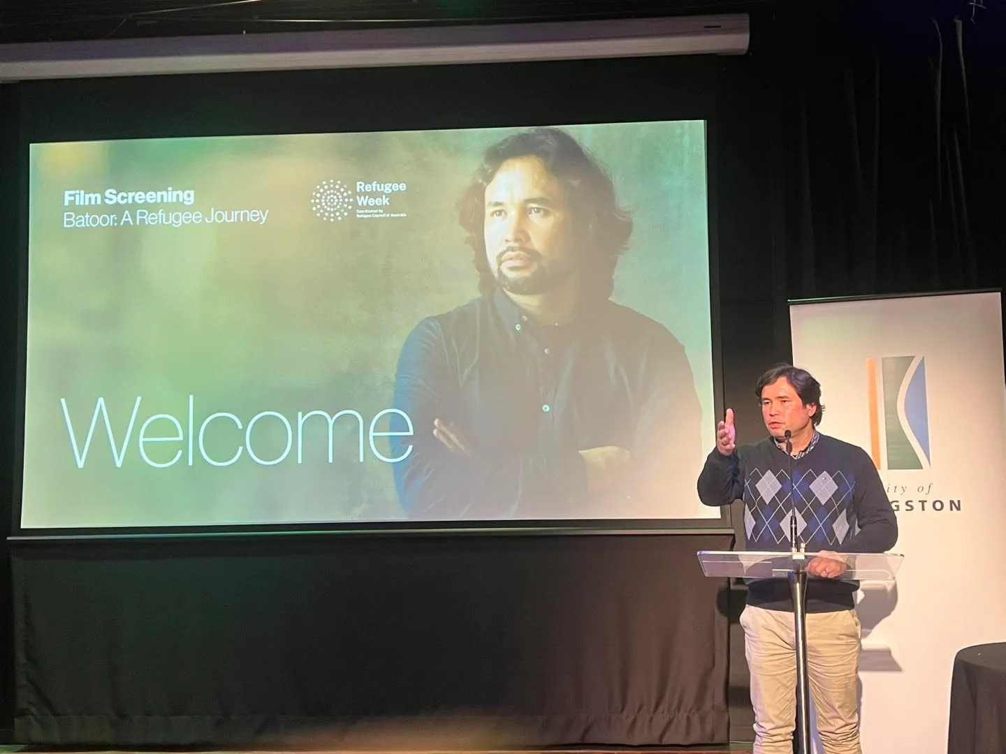 Barat Ali Batoor at a lectern at a Refugee Week screening of Batoor: A Refugee Journey in Kingston, his portrait on the screen behind him.