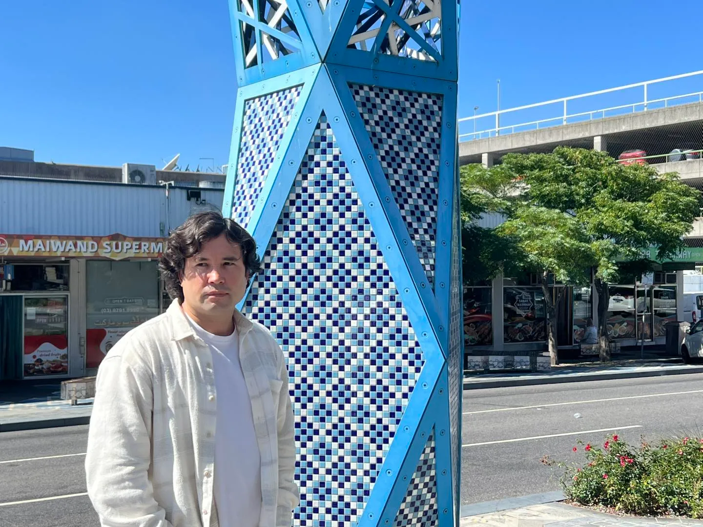 Barat Ali Batoor beside a tall blue-tiled tower on a Dandenong street, shopfronts behind him.